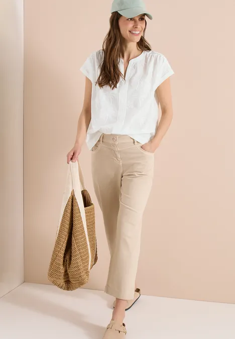 Vrouw in witte blouse en beige broek met strooien tas, voor een lichtbeige achtergrond.