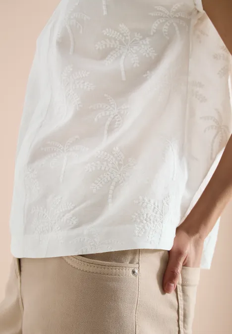 Close-up van een persoon in een witte, geborduurde top met bladmotief en beige broek.
