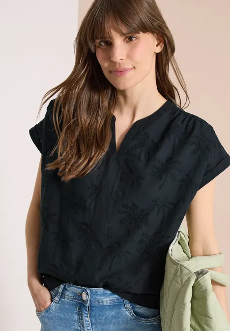 Vrouw draagt zwarte, kort mouwen blouse met subtiel patroon, spijkerbroek en houdt groene jas over haar arm.