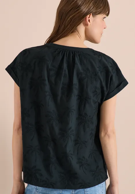 Vrouw van achteren in een losse zwarte top met korte mouwen en subtiel borduursel met bladmotief.