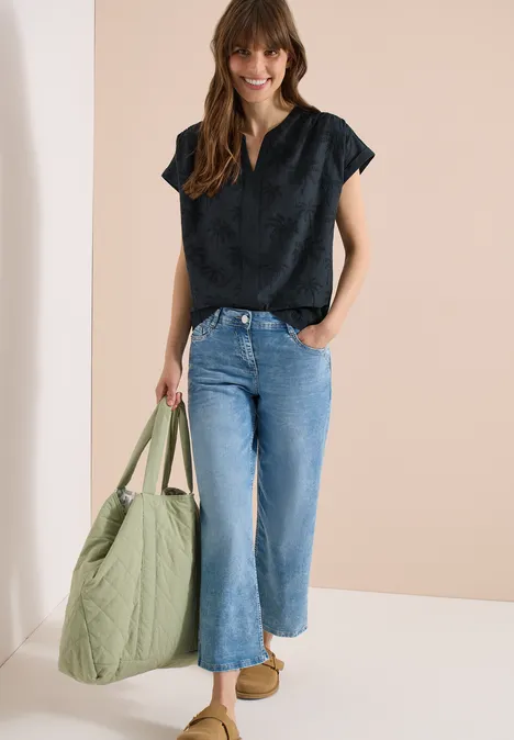 Vrouw in zwart topje en wijde blauwe jeans, met grote olijfgroene tas en beige schoenen.