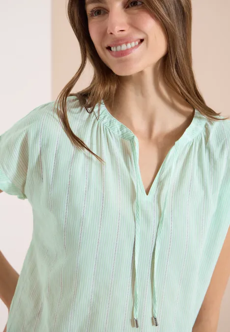 Frau trägt mintgrüne, kurzärmlige Bluse mit feinen weißen Streifen und V-Ausschnitt mit Kordel.