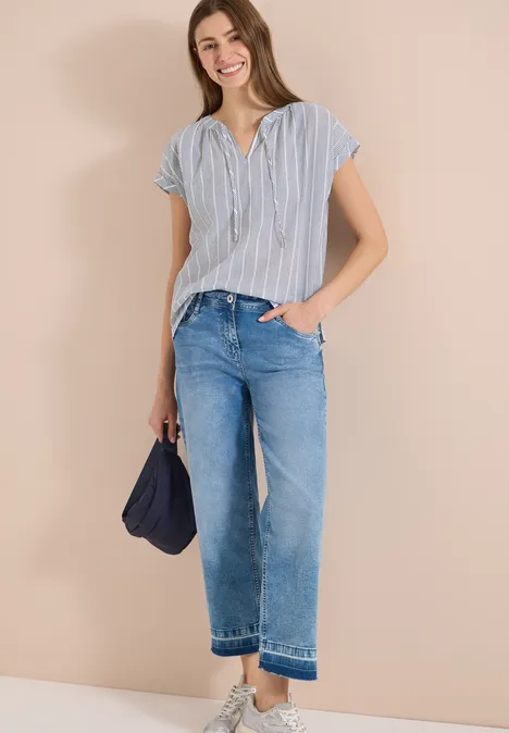 Dame in gestreiftem Kurzarmhemd, blauen Jeans und dunkelblauer Tasche vor beigem Hintergrund.