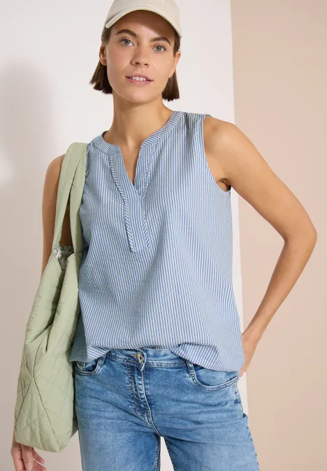 Vrouw draagt lichtblauw mouwloos shirt, spijkerbroek, groene tas op schouder en witte pet.