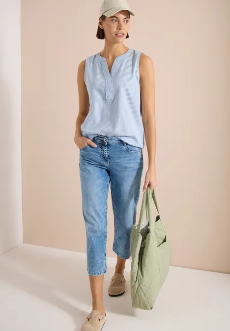 Vrouw in lichtblauwe mouwloze top, spijkerbroek en espadrilles met een grote groene tas voor een neutrale achtergrond.