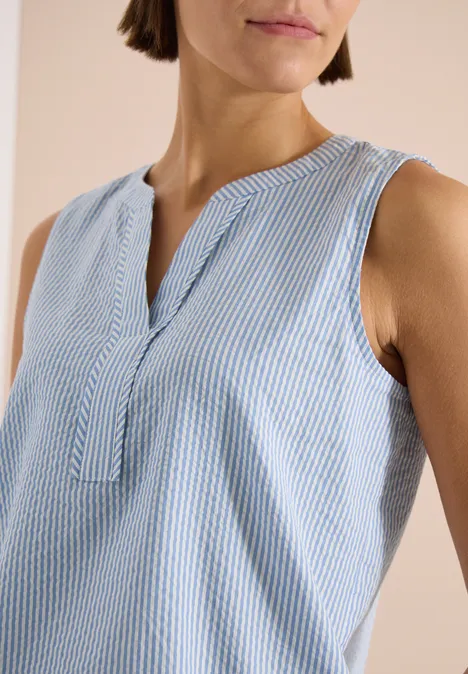 Vrouw in mouwloze, blauw-wit gestreepte top met splitkraag tegen lichte achtergrond.