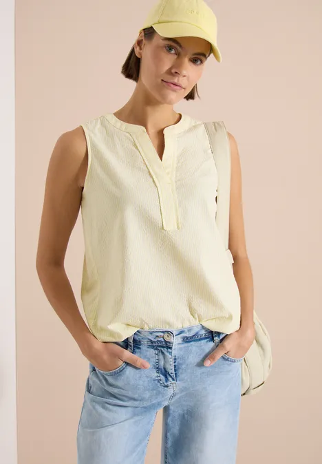 Personne portant un top sans manches jaune pâle et un jean bleu clair, mains dans les poches, fond rose.
