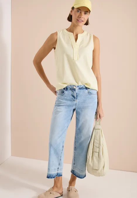 Femme en top sans manches crème, jean bleu clair et sac assorti devant fond neutre.
