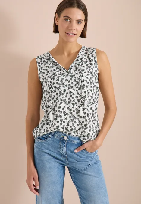 Vrouw in mouwloze witte top met groen motief en V-hals, gecombineerd met een blauwe spijkerbroek.