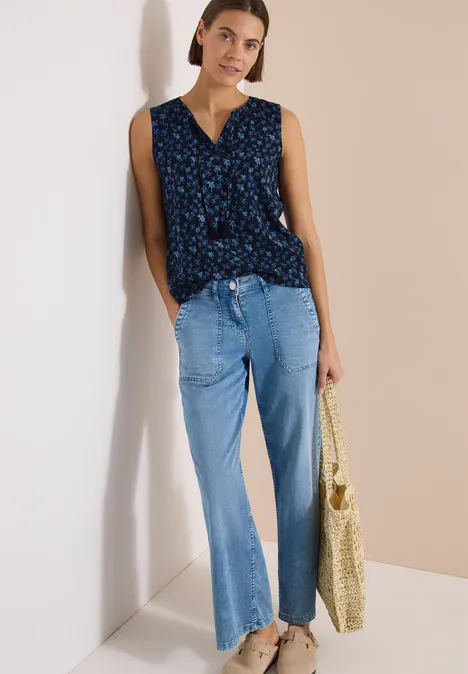 Vrouw in mouwloze blauwe bloemen-top, lichte jeans en een strooitas voor een minimalistische tweekleurige achtergrond.