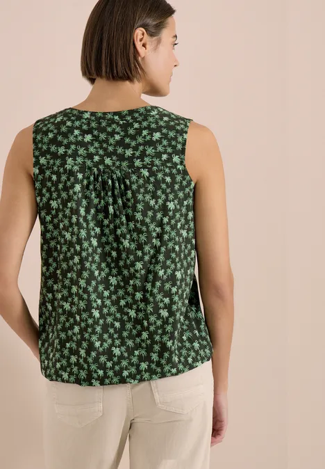 Person mit kurzem Haar von hinten: trägt ein ärmelloses grünes Oberteil mit weißem Blumenmuster und helle Hose.