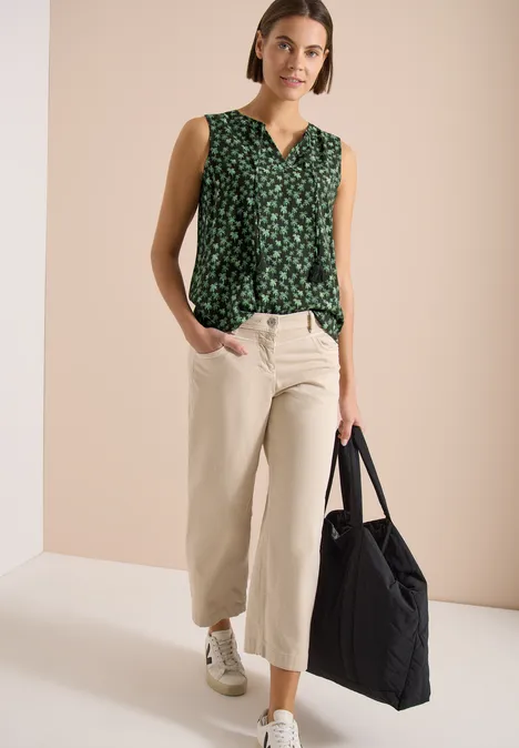 Frau trägt ärmellose grüne Bluse mit Blumenmuster, beige weite Hose, weiße Sneakers und schwarze Tragetasche.