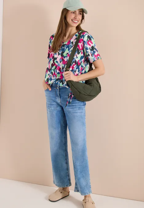 Frau trägt bunte Bluse, gerade geschnittene Jeans und grüne Umhängetasche vor beigem Hintergrund.
