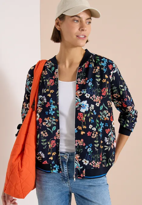 Eine Frau trägt eine schwarze Bomberjacke mit buntem Blumenmuster, Jeans, weißes Top, Cap und orangefarbene Tasche.