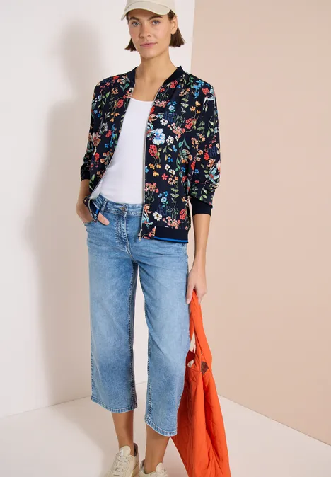 Frau trägt florale Bomberjacke, weißes Shirt, ausgestellte Cropped-Jeans und eine orangefarbene Tasche.