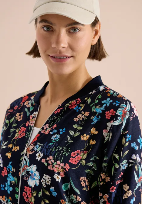 Person trägt dunkelblaue Bomberjacke mit buntem Blumenmuster und weiße Baseballkappe