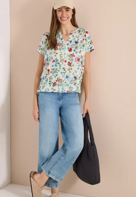 Vrouw draagt een kleurrijke bloes met bloemenprint, wijde blauwe jeans, sandalen en een zwarte tas voor een lichte achtergrond.