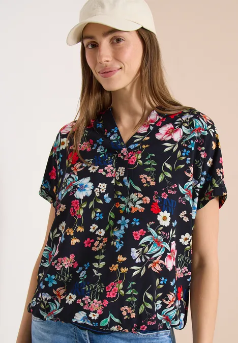 Vrouw tegen lichte achtergrond draagt zwarte bloemenblouse en beige pet.