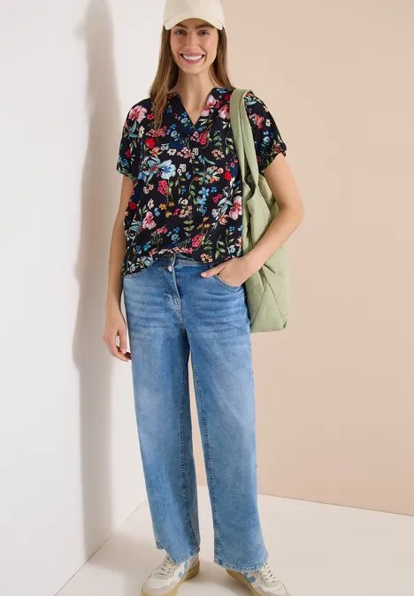 Vrouw met zwarte bloemenblouse, lichtblauwe wijde jeans en groene shopper.