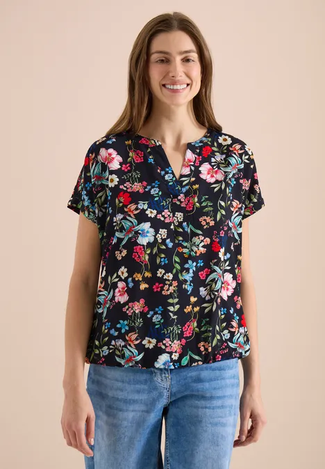 Vrouw in zwarte bloes met gekleurd bloemenpatroon en blauwe jeans, van voren gefotografeerd.