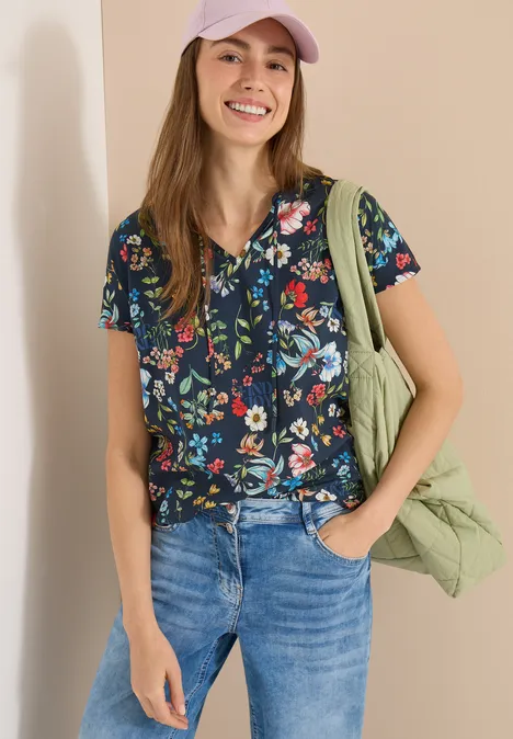 Frau in Jeans, dunkelblauer Bluse mit bunten Blumen, grünem Schulterbeutel, eine Hand in der Hosentasche.