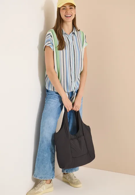 Hemdbluse mit Streifen und Knoten light sky blue Frau trägt gestreiftes Hemd, weite Jeans, Sneaker und hält eine schwarze Tasche vor beigem Hintergrund.
