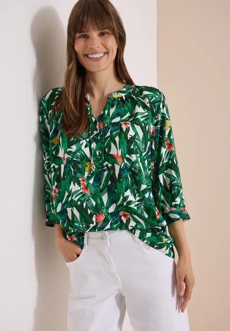 3/4-Arm Tunika mit Split Neck und Print khaki Frau in grüner, tropischer Bluse mit Papageienmuster und weißen Hosen vor neutralem Hintergrund.