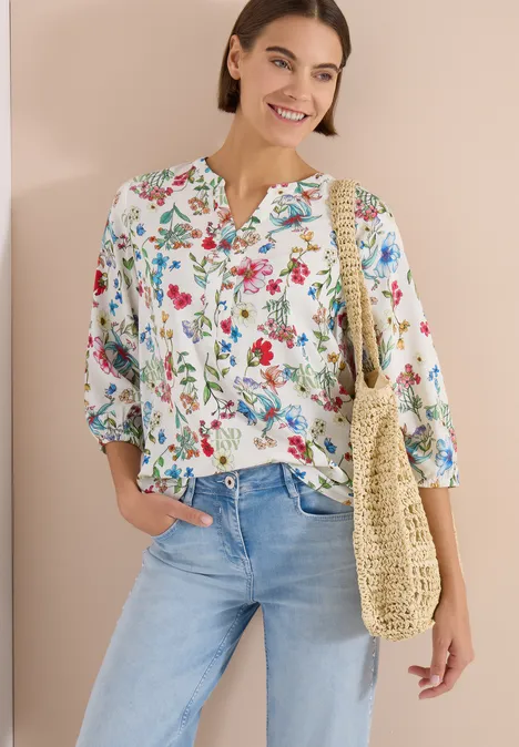 Frau in floraler Bluse, hellen Jeans und sommerlicher Stroh-Netztasche.