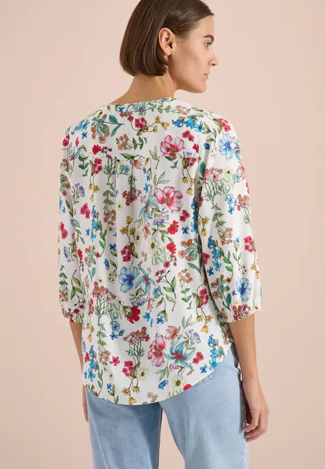 Frau von hinten in weißer Bluse mit bunten Blumen und Jeans vor hellem Hintergrund.