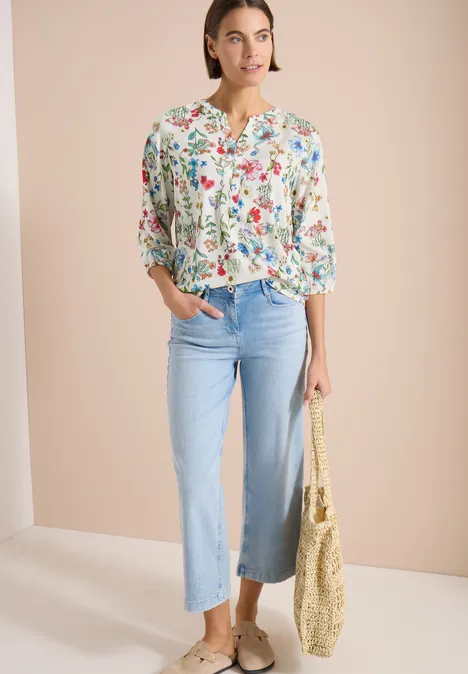Frau mit geblümter Bluse, hellblauen weiten Jeans, Espadrilles und Strohtasche vor hellem Hintergrund.