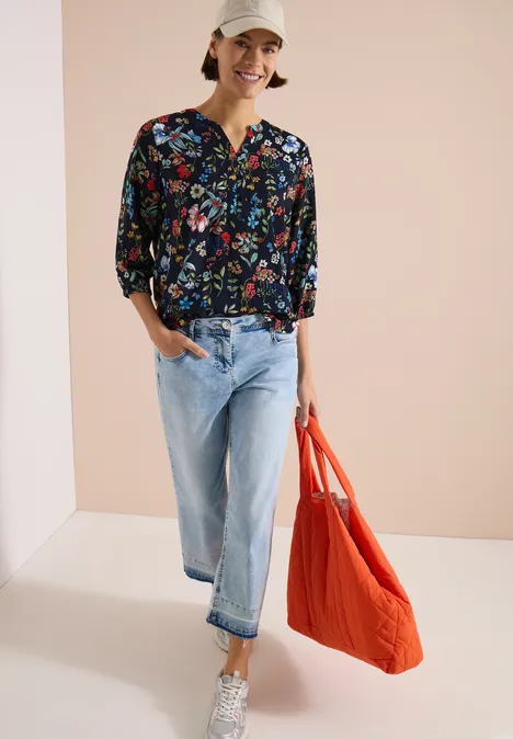 3/4-Arm Bluse mit Split Neck und Print universal blue Dame in geblümter Bluse, blauen Jeans, weißen Sneakern und orangefarbener Tasche vor hellem Hintergrund.
