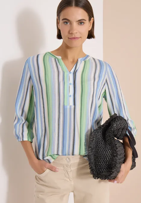 Frau trägt bunte, gestreifte Bluse mit 3/4-Ärmeln, beige Hose und hält eine schwarze Flecht-Tasche.