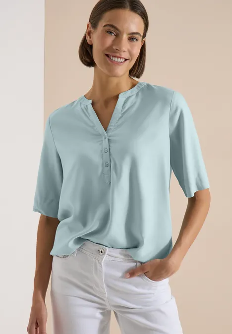 Blouse met korte mouwen en gespleten hals crystal mint Eine Frau in einer hellblauen Bluse mit halblangen Ärmeln und weißen Hosen, eine Hand in der Tasche.
