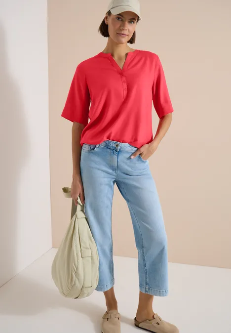Blouse met korte mouwen en gespleten hals coral Persoon draagt rood poloshirt, lichtblauwe jeans en beige tas.