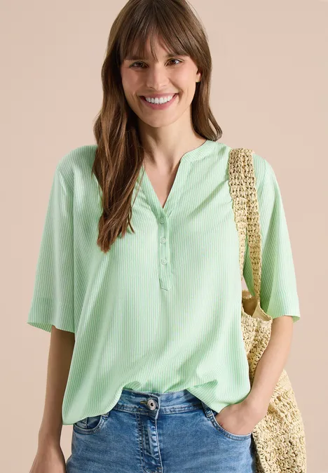Frau in pastellgrünem Kurzarmhemd mit Knopfleiste, blauen Jeans und Strohtasche vor beigem Hintergrund.