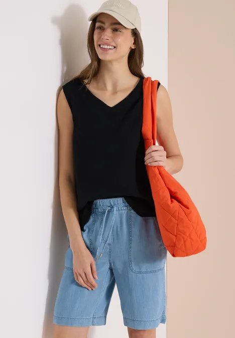 Person in ärmellosem schwarzen V-Ausschnitt-Top, blauen Jeans-Shorts, Cap und orangener Stepp-Umhängetasche