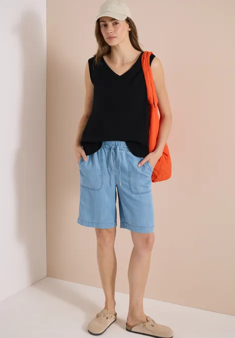 Eine Frau in schwarzem ärmellosem Top, hellblauen Jeans-Bermudas und beigen Loafers mit orangefarbener Tasche.
