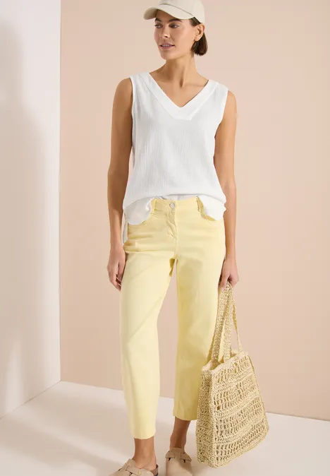 Frau trägt weißes ärmelloses V-Shirt, gelbe Hose und einen gehäkelten Strohtasche.