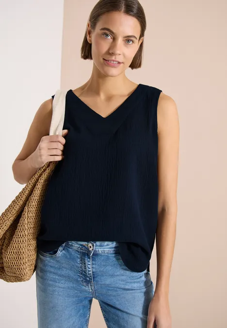 Frau im ärmellosen dunkelblauen V-Ausschnitt-Top, Jeans und Strohtasche vor hellem Hintergrund.
