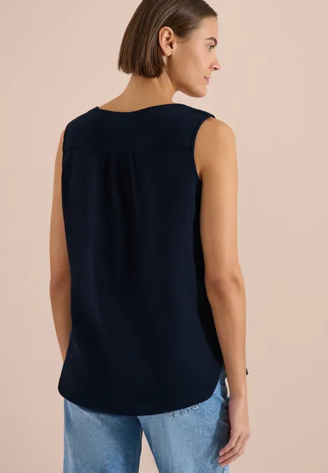 Frau von hinten in ärmellosem dunkelblauem Top und hellblauen Jeans vor neutralem Hintergrund.