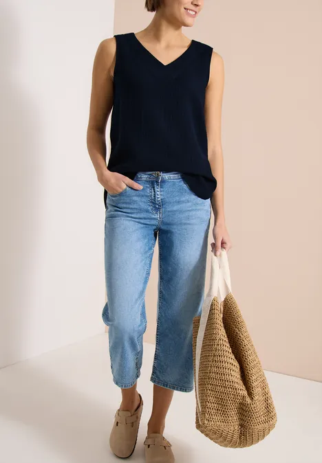 Frau in schwarzem, ärmellosem V-Ausschnitt-Top und lockeren, hellblauen Jeans mit großer Basttasche.