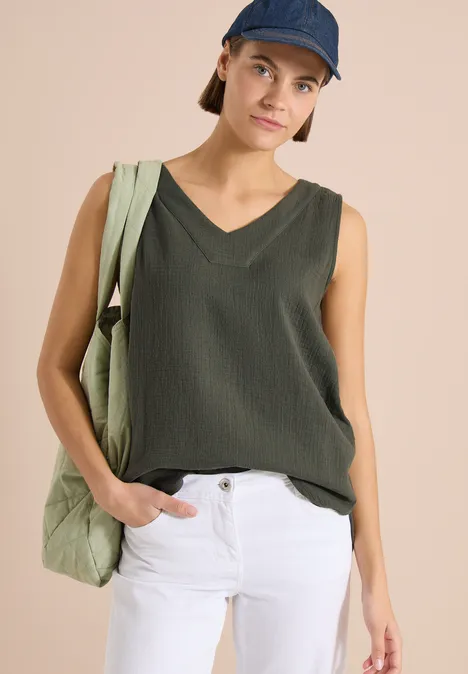 Frau in ärmellosem Oliv-Top, weißer Hose und Cap, trägt lässig eine Stofftasche.
