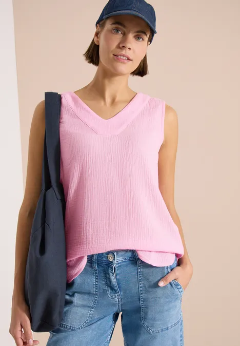 Person trägt rosa ärmelloses Top, blaue Jeans, blaue Tasche und Cap vor schlichtem Hintergrund.