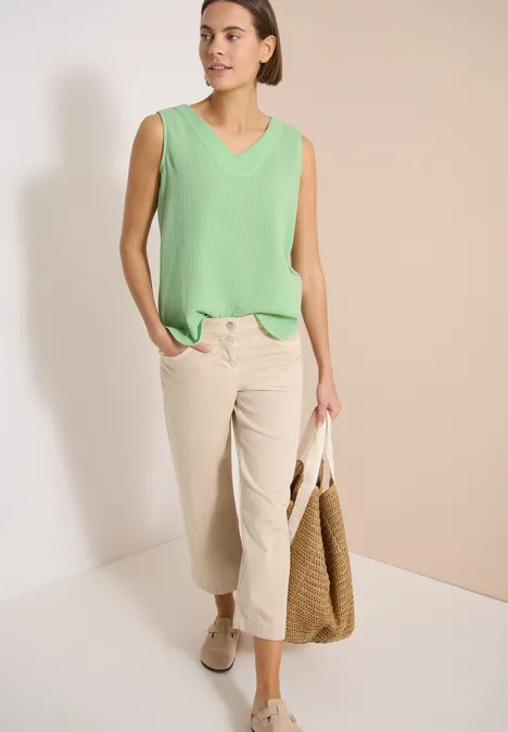 Frau in ärmellosem mintgrünen Top, cremefarbener Culotte, beigen Slippern und Strohtasche vor neutralem Hintergrund.
