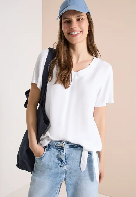 Frau trägt weiße, locker sitzende Bluse mit Knotendetail, blaue Jeans und schwarzen Schulterbeutel vor hellem Hintergrund.