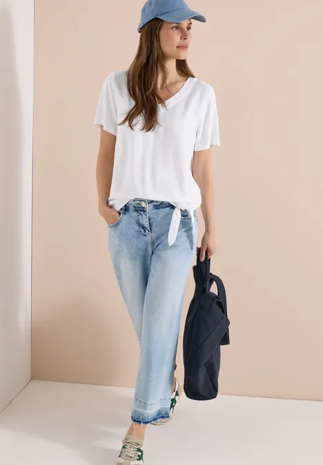 Frau in weißem T-Shirt, hellblauen Jeans, Cap und Sneakers hält eine dunkelblaue Tasche.