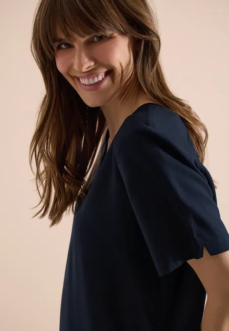 Seitliches Porträt einer Frau in dunkelblauem, kurzärmligem Oberteil vor hellem Hintergrund.