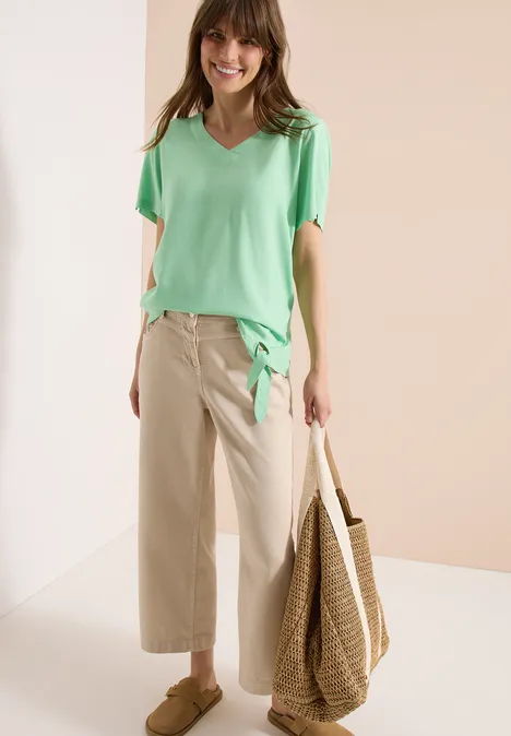 Model in lichtgroen V-shirt, beige wijde broek en grote strooien tas in minimalistische setting.