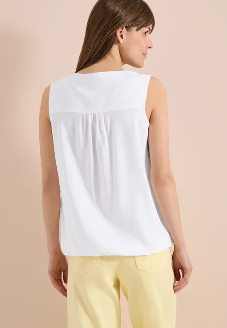 Frau von hinten in weißem ärmellosem Oberteil mit Falten und gelben Hosen vor beigem Hintergrund.