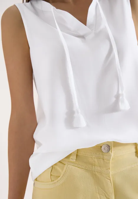 Nahaufnahme einer Person mit ärmellosem weißen Kapuzen-Top mit Kordeln vorne und gelben High-Waist-Hosen.